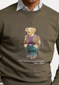 Suéter verde oliva con un oso bordado que lleva una chaqueta de tweed, suéter morado y pantalones de rayas verdes, completado con zapatos marrones.