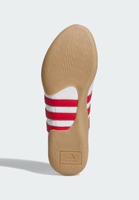 Potplat tenisice marke Adidas, s bazom od smeđe gume, bijelim gornjim dijelom i tri crvene pruge. Uključuje teksturirani uzorak za bolje prianjanje.