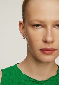 Pendiente de aro de plata con acentos puntiagudos. El modelo lleva puesta una blusa texturizada verde, que presenta un escote redondo y un dobladillo asimétrico.