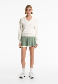 Witte gebreide trui met een V-hals en geribbelde zoom, gecombineerd met een groene uitlopende rok met een witte rand, gedragen met witte sportschoenen.