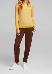 Gelber Strickpullover mit Wabenmuster, kombiniert mit braunen, schmal zulaufenden Hosen und weißen Sneakers, die umgeschlagene Ärmel und ein schlichtes Design aufweisen.