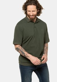Olijfgroene polo met een knoopsluiting bij de kraag, korte mouwen en een subtiele logo. Gepaard met vervaagde blauwe jeans, waar gedetailleerde arm tatoeages zichtbaar zijn.
