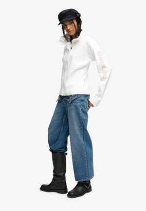 Kvinde iført en hvid lynlåstrøje med teksten "Bench" på ærmet, løse blå jeans, sorte støvler og en sort kasket, stående i en afslappet positur.