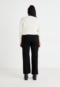 Cremig-gestreifter Pullover kombiniert mit hochtaillierten schwarzen Jeans. Schwarze Blockabsatz-Stiefeletten vervollständigen das Outfit. Einfarbiger Hintergrund.