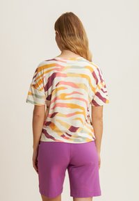 T-shirt a maniche corte di colore crema con motivi a onda multicolore, abbinato a pantaloni corti viola. Il tessuto sembra morbido e ha una vestibilità rilassata.