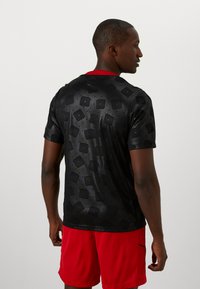 Camisa negra de manga corta con una textura estampada que presenta cuadrados y puntos. Detalle en el cuello rojo. Tela ligera y suave.