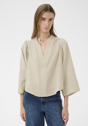 Kobieta w beżowej bluzce w prążki z szerokimi rękawami i niebieskich dżinsach, stojąca na tle jasnego, jednolitego tła.