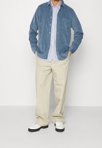 Mans blårandig kordflor skjorta över en vit randig skjorta, krämfärgade byxor och vita loafers med svarta sulor.