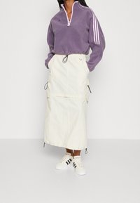 Lila fleece halvdraget tröja med rosa dragkedja och tre vita ränder; krämfärgad maxikjol med cargofickor och justerbara snören; vita sneakers.