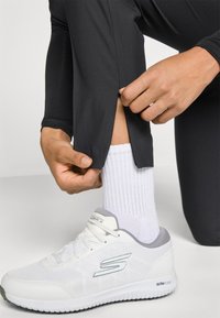 Pessoa a ajustar o fecho de correr de umas calças de treino pretas, acima de um tênis branco com detalhes cinzentos e meias brancas caneladas.