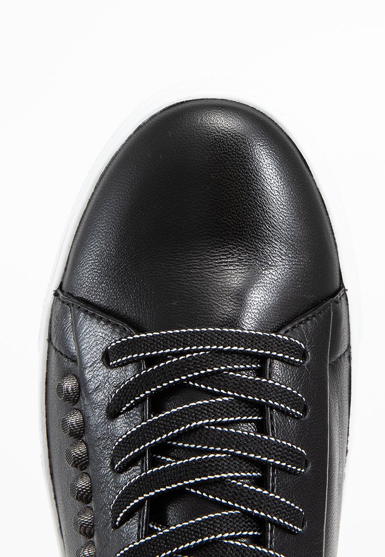 Schwarze Leder-Sneaker mit schwarz-weißen Akzenten an den Schnürsenkeln und dunklen Metallnieten an der Seite, vor einem weißen Hintergrund.