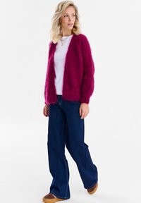 Blondynka w puszystym fioletowym kardiganie na białej koszuli, ciemnych szerokich dżinsach i brązowych butach, patrząca w bok.