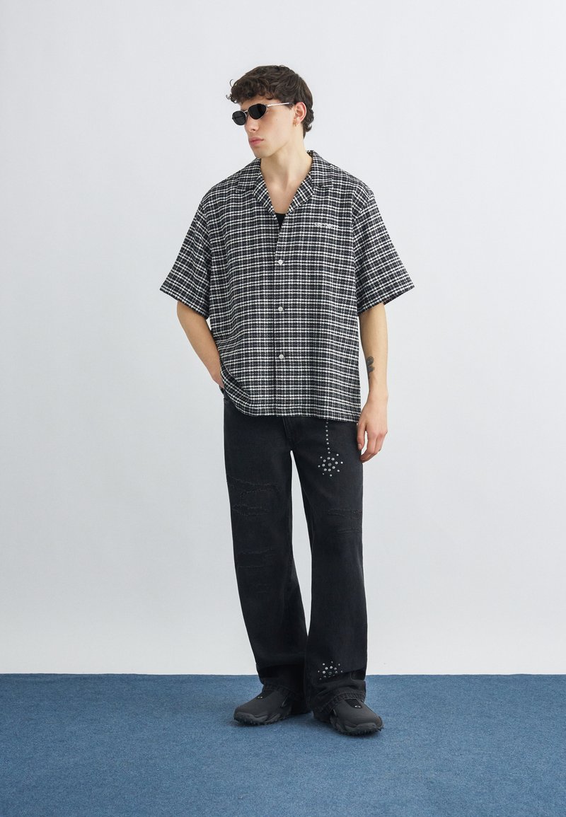 Giovane uomo che indossa occhiali da sole neri, camicia a maniche corte a quadri bianco e nero, pantaloni neri con motivi decorativi e scarpe nere, in piedi.