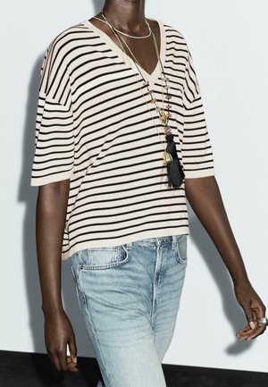 Person med mørk hud iført en creme- og sortstribet V-hals top, lyseblå jeans og lagdelte halskæder med vedhæng og frynser.