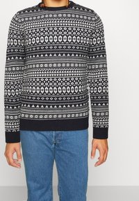 Pull bleu marine et blanc à motifs avec un col rond, mettant en avant des dessins géométriques et floraux, avec des poignets et un ourlet côtelés, porté avec un jean bleu.