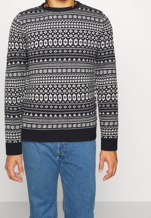 Pull bleu marine et blanc à motifs avec un col rond, mettant en avant des dessins géométriques et floraux, avec des poignets et un ourlet côtelés, porté avec un jean bleu.