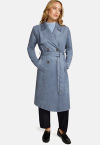 Trench-coat en daim bleu clair avec des boutons croisés, une ceinture nouée et des poches latérales. Porté sur un pull bleu pâle et un pantalon foncé.