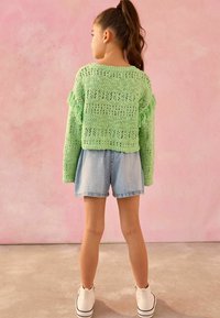 Grüner gestrickter Pullover mit Fransen, kombiniert mit hellblauen Denim-Shorts und weißen High-Top-Sneakern. Der Hintergrund ist pink.