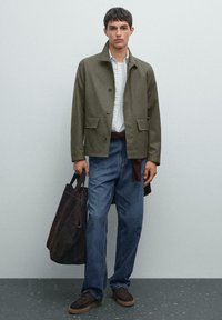 Khaki jakke med frontknapper og to påsætningslommer, parret med blå jeans. Modellen bærer en mørkebrune tote bag. Brune sko fuldender outfittet.