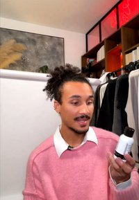 Personne en pull rose tenant et examinant une bouteille blanche dans une pièce avec des portant à vêtements et des étagères à livres.