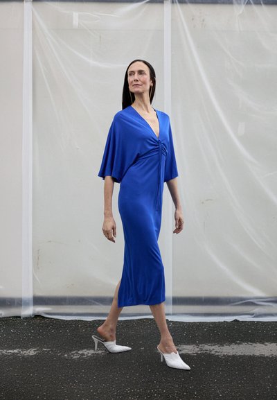 Modra midi obleka z nabrano zasnovo in V-izrezom, z vozelnim detajlom spredaj. Oblečena z belimi mule natikači z visokimi petami.