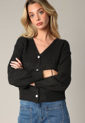 Cardigan noir en maille avec un col en V, présentant un motif texturé et trois boutons blancs sur le devant. Longueur courte avec des manches longues.