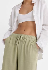 Nainen, jolla on päällään valkoinen crop-top ja auki oleva valkoinen nappipaita, kevyet vihreät nyöritaskuhousut, kädet taskuissa.