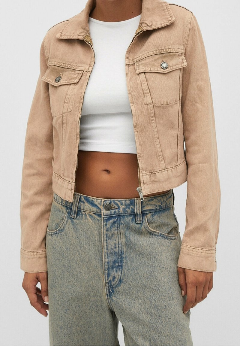 Personne portant une veste en jean beige courte par-dessus un top blanc court et un jean bleu clair taille haute.