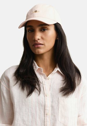 Woman with long dark hair wearing a pale pink button-up shirt and light pink cap with small embroidered logo, neutral background.