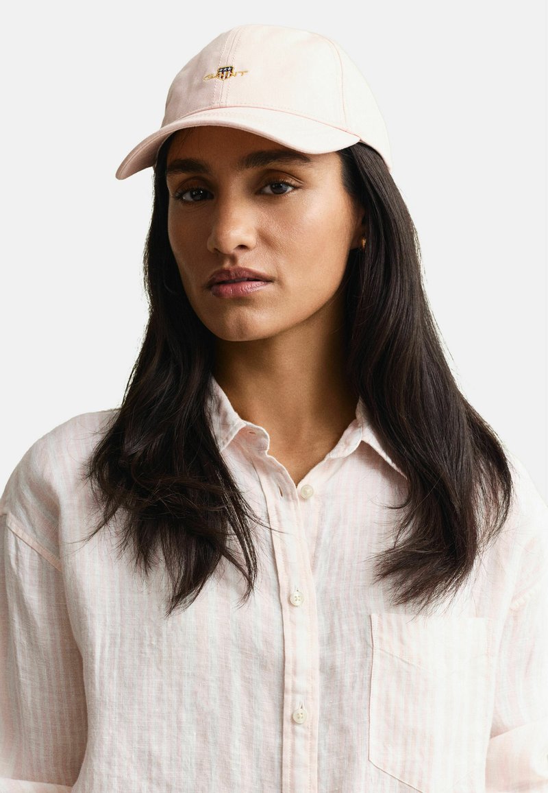 Femme aux longs cheveux foncés portant une chemise à boutons rose pâle et une casquette rose clair avec un petit logo brodé, fond neutre.