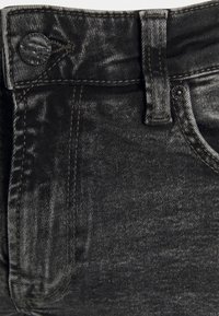 Primer plano de unos jeans de mezclilla negra mostrando la cintura, el pasador del cinturón, el botón y un bolsillo con detalles de costura en contraste.