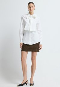 Witte blouse met grote strik bij de kraag, gecombineerd met een bruine gestructureerde rok. Zwarte slingback schoenen met gouden accenten maken de look compleet.