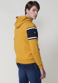 Un modello in una felpa gialla senape con strisce blu e cappuccio, rivolto leggermente all'indietro, abbinato a jeans blu, su uno sfondo chiaro.
