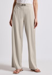 Pantalons beiges à jambes larges avec une taille smock�ée et un détail de boucle en métal. Texture lisse, design fluide, coupe ajustée avec des coutures frontales.