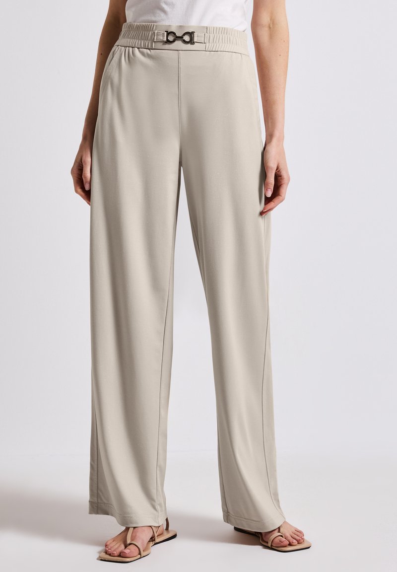 Pantalons beiges à jambes larges avec une taille smockée et un détail de boucle en métal. Texture lisse, design fluide, coupe ajustée avec des coutures frontales.