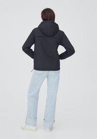 Chaqueta negra con capucha y corte recto, hecha de material suave. Combinada con jeans de pierna ancha de color azul claro y zapatos blancos.