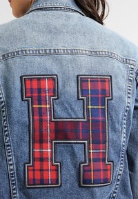 Jeansjacka med en stor broderad rutig bokstav "H" i röd, blå och gul på baksidan. Texturen är jämn med synliga stygn.