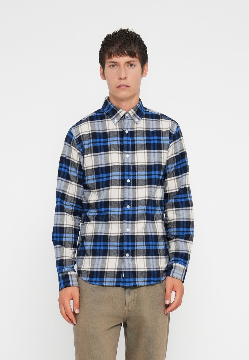 Camisa de botão em padrão xadrez azul e cinza, com mangas longas, feita de um tecido macio; apresenta um colarinho clássico e botões brancos. Usada com calças caquis.