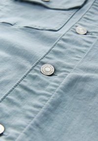 Light blue shirt fabric with visible texture, featuring silver snap buttons and a pocket, showing stitching and softness.