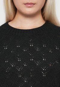 Melnā adītā džemperī ir strukturēts raksts ar dimanta formām, tam ir atvērtas detaļas un ribota apkakle.