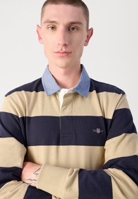 Beige és tengerészkék csíkozott hosszú ujjú pólóing világoskék gallérral és kis logóval a mellen. Az anyag lágy hatásúnak tűnik, textúrált felülettel.