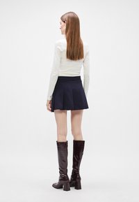 Λευκή μπλούζα με μακριά μανίκια, ναυτική πλισέ μίνι φούστα και μποτάκια μέχρι το γόνατο σε μπορντό χρώμα με υφή και χοντρό τακούνι. Μοντέλο από πίσω.