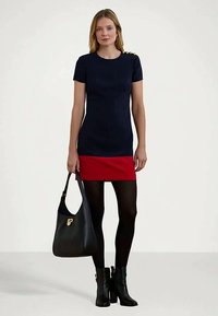 Mulher vestindo um vestido de manga curta azul-marinho e vermelho, meias-calças pretas, botins pretos e segurando uma bolsa preta com fecho dourado.