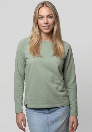 Junge Frau mit langem blondem Haar, die ein hellgrünes Langarmshirt und einen hellblauen Jeansrock trägt, lächelnd vor einem schlichten Hintergrund.