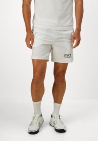 Muškarac u bijelim sportskim šortsevima EA7 Emporio Armani, bijelim čarapama i bijelim atletskim cipelama s crnim i žutim detaljima stoji na bijelom podu.
