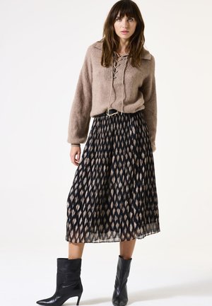 Un pull beige en maille avec un détail à lacets, associé à une jupe plissée noire ornée d'un motif géométrique crème. Bottines noires.