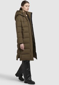 Polstret lang frakke i olivengrøn med hætte, der har en lynlås, sidelommer og en glat tekstur. Båret over sorte læderbukser og støvler.