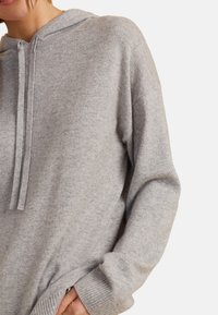 Helle graue Kapuzenjacke aus weichem Strickmaterial. Verfügt über einen Kordelzug, gerippte Manschetten und eine lockere, oversized Silhouette.