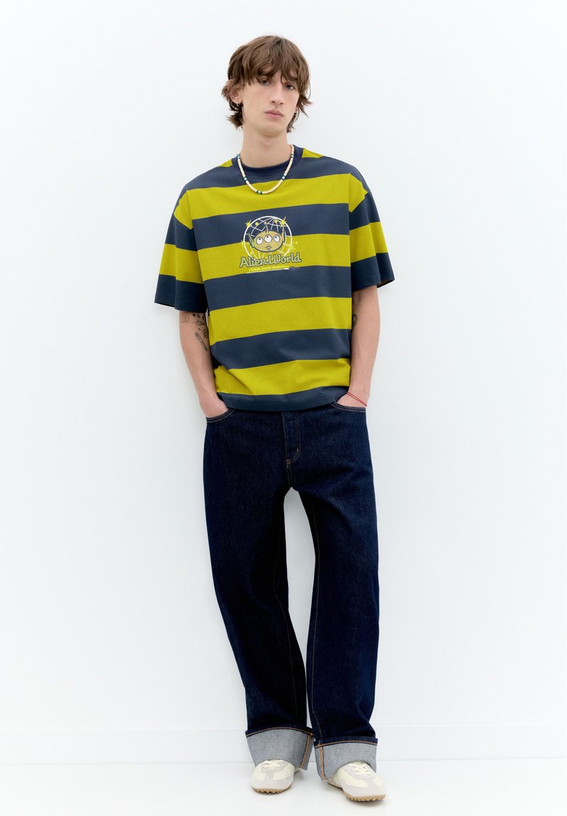 Jeune homme debout contre un mur blanc portant un t-shirt rayé bleu foncé et jaune avec un motif d’extraterrestre, un jean large foncé, des baskets blanches et un collier de perles.