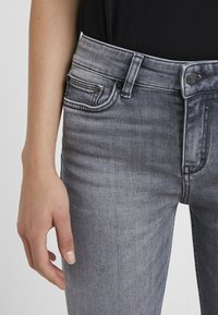Jeans de mezclilla gris con un acabado desgastado, diseño de cinco bolsillos y detalles en metal negro; la tela tiene una textura suave.